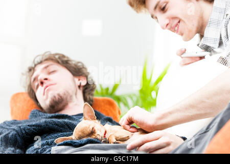 Sorridente giovane uomo petting long eared chihuahua cane addormentato sulla parte superiore del torace di stanco fratello gemello Foto Stock