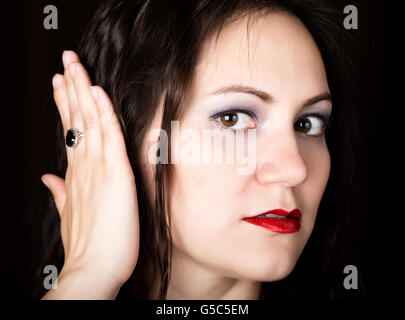 Close-up donna guarda dritto verso la fotocamera su uno sfondo nero. ridendo donna copre le sue orecchie con le sue mani. esprime emozioni diverse Foto Stock