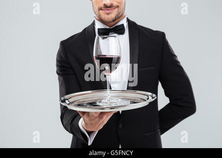 Primo piano della giovane maggiordomo in smoking tenendo il vassoio in argento con un bicchiere di vino rosso Foto Stock