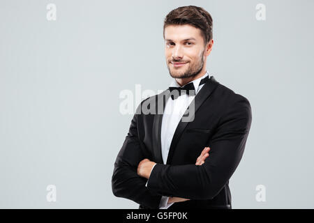 Sorridente fiducioso giovane uomo in smoking in piedi con le braccia incrociate su sfondo bianco Foto Stock