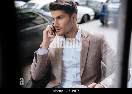 Attraente giovane imprenditore seduto e parlando al telefono cellulare seduti all'aperto in cafe Foto Stock