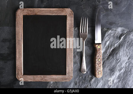 Vista dall'alto di un coltello e forchetta accanto a un menu vuoto lavagna. Una forte luce laterale su un nero ardesia tabella. Foto Stock