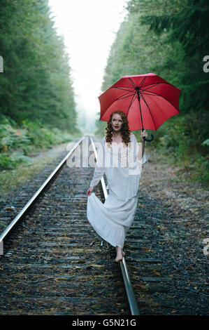 La donna caucasica camminando sui binari del treno con ombrellone in foresta Foto Stock