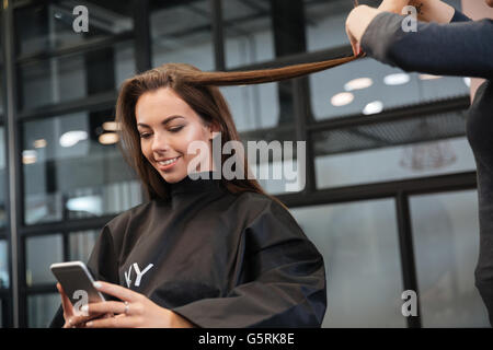 Felice giovane donna con lo smartphone e parrucchiere rendendo lo styling dei capelli a salon Foto Stock