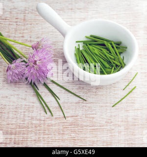 Overhead luminoso cresciuto in casa erba cipollina tritata in un piatto bianco con fiori viola, la finestra illuminata per messa a fuoco morbido, legno sfondo della granella Foto Stock