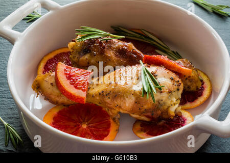 Pollo arrosto gambe su fette di arance rosse in bianco teglia. Nero ardesia sfondo. Foto Stock