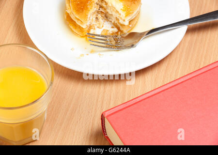 Pila di parte mangiato frittelle ricoperto di sciroppo su un tavolo per la colazione con un libro e un bicchiere di succo di arancia Foto Stock