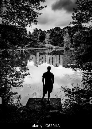 Silhouette di uomo si trovava su un molo di legno che si affacciava sul lago con gli alberi che circondano i bordi Foto Stock