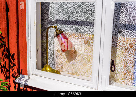 Bella vintage lampada da tavolo in una vecchia finestra con pizzo bianco dietro a tendina. È un edificio a red cottage di legno con finestra bianca Foto Stock