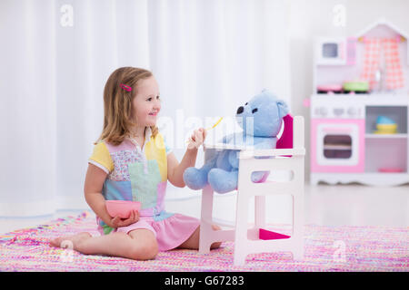 Poco carino ragazza che gioca con il suo orsacchiotto di peluche. Alimentazione bambino bambola in vivaio bianco. I bambini giocano con legno giocattoli educativi. Foto Stock