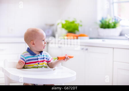 Happy Baby sitting in alta sedia mangiando la carota in una cucina bianca. Una sana alimentazione per i bambini. Foto Stock