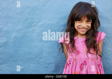 Ritratto di carino bambina guardando la fotocamera e sorridere mentre in piedi contro la parete di blu. Innocenzo giovane femmina bambino con co Foto Stock