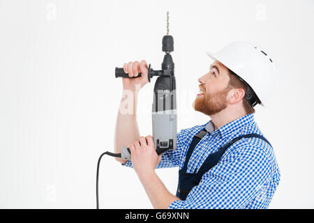 Sorridente giovane barbuto builder nel casco mediante trapano e guardando in alto Foto Stock