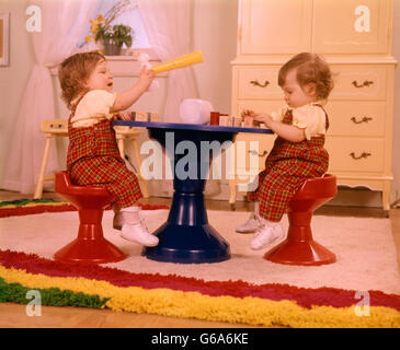 Anni Settanta due letti i bimbi che indossa RED PLAID tuta seduti su sgabelli in plastica blu tabella giocando con i giocattoli Foto Stock