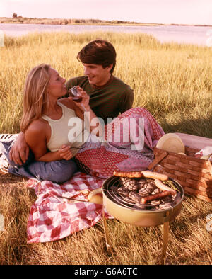 Anni Settanta coppia giovane PICNIC IN ERBA bevendo vino hot dogs sul grill Foto Stock