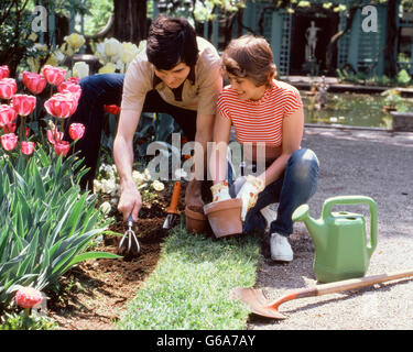 Anni ottanta GIOVANE UOMO GIARDINAGGIO SCAVARE NEL LETTO DI FIORI donna inginocchiato accanto a lui con vasi da fiori ANNAFFIATOIO E PALA Foto Stock