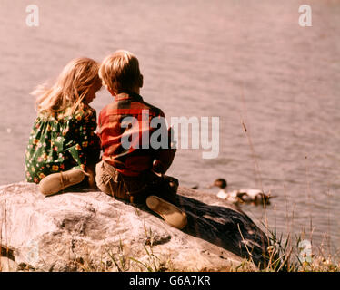 Anni Settanta due bambini ragazzo ragazza seduta insieme su una roccia affacciato su un lago di anatre di alimentazione Foto Stock