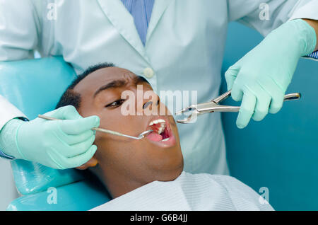 Giovane uomo ispanico giacente in cattedra riceve il trattamento dentale con la bocca aperta, dentista mani indossando i guanti azienda utensili per la lavorazione su pazienti denti Foto Stock