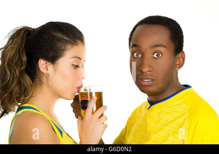 Affascinante coppia interraziale giallo da indossare magliette da calcio, posa per fotocamera mentre la donna beve dal bicchiere di birra e di uomo con sorpresa l'espressione del viso, bianco di sfondo per studio Foto Stock