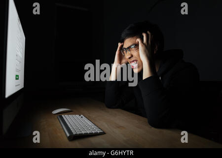 Mad irritato asian giovane uomo che lavora con il computer nella stanza buia Foto Stock