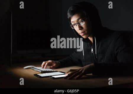 Considerato giovane utilizzando il computer, schermo bianco tablet e cellulare in camera oscura Foto Stock