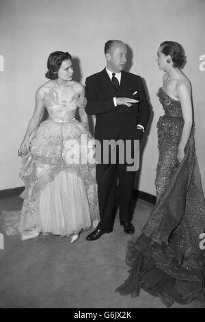 Il famoso designer francese Christian Dior, esteso a mano, sottolinea un punto di moda mentre parla con due dei suoi modelli alla sfilata di moda del Savoy Hotel di Londra. L'abito sulla sinistra è intitolato 'Mozart' ed è il vestito più costoso dello spettacolo. E' di tulle di champagne pallido, ricamato in diamante e paglia. L'abito a destra è il vestito di gala 'Schumann' in tulle bianco, rifinito con pizzo valenciano. Foto Stock