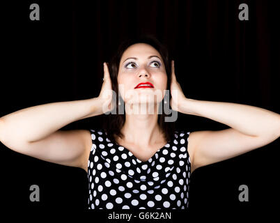 Close-up donna guarda dritto verso la fotocamera su uno sfondo nero. ridendo donna copre le sue orecchie con le sue mani. esprime emozioni diverse Foto Stock