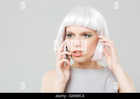 Riflessivo preoccupato giovane donna in parrucca bionda parlando al telefono cellulare su sfondo bianco Foto Stock