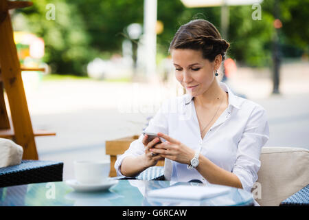 Ritratto di un affascinante business donna in chat sul suo smart phone mentre si è in attesa di qualcuno in cafe' all'aperto, bellissima femmina utilizzando il telefono cellulare mentre è seduto in un accogliente ristorante nella fresca aria durante il pranzo Foto Stock