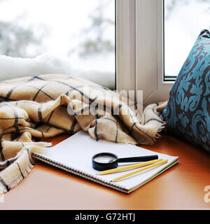 Aprire il blocco note, vetro magnifier, cuscino, matite e beige caldo plaid situato sul legno stilizzata davanzale. Concetto di inverno di com Foto Stock