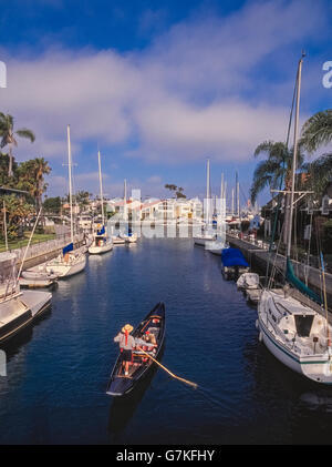 Una paglia-cappello gondoliere utilizza un unico remo alla paletta di un tradizionale gondola veneziana e prendere una giovane coppia romantica su una rilassante crociera attraverso i canali di Napoli isola vicino alla riva di Belmont area di Long Beach, California, Stati Uniti d'America. Uno studente universitario che ha iniziato la fuga in Gondola company nel 1982 ora dispone di una flotta di dieci gondole e offre tour giornalieri durante il giorno e dopo il tramonto. Foto Stock