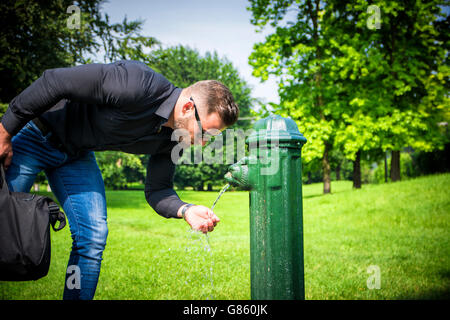 Vista laterale della trafficata uomo in occhiali da sole con la valigia di bere acqua dalla fontana nel parco di estate Foto Stock