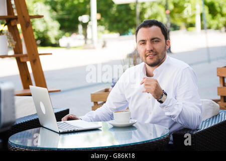 Giovane uomo bello sorridente e di bere il caffè in un ristorante sul mare. Egli è felice, guardando la fotocamera Foto Stock