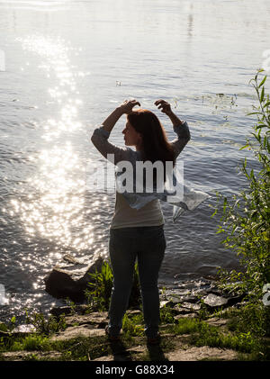 Donna in piedi dal fiume con le braccia sollevate e sciarpa soffiando nel vento Foto Stock