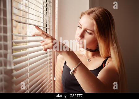 Ragazza triste guardando nella finestra attraverso le persiane Foto Stock