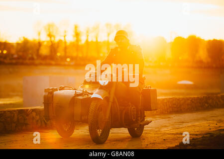 Unidentified ri-enactor vestita come la II Guerra Mondiale il tedesco soldato della Wehrmacht che corre sul motociclo in raggi del sole al tramonto. Foto Stock