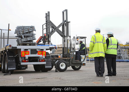 Un camion-montato carrello elevatore scarica ponteggi attrezzature da un pianale, autoarticolato rimorchio, guardato da due uomini Foto Stock