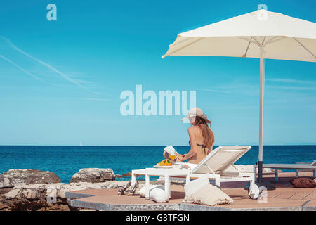 Donna con cappello per il sole rilassandovi sulla terrazza sulla spiaggia Foto Stock