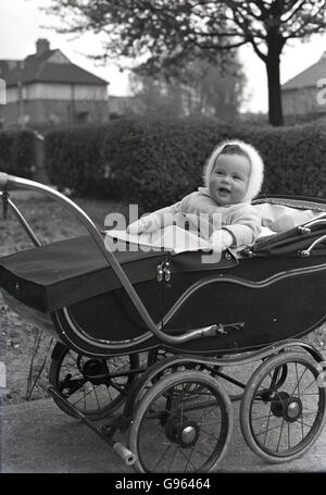 anni '50, quadro storico, un bambino seduto in su in una tradizionale croce d'argento carrozza costruita su un percorso all'esterno in un giardino anteriore. Queste eleganti ed eleganti carrozzine avevano un telaio largo e ruote in filo metallico e con un telaio robusto e un materasso imbottito significavano una comoda corsa per il bambino. Foto Stock