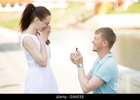 Bel giovane la richiesta di mano d'amata bella ragazza, chiedendo di sposarsi. Sorridente guy standing sul suo ginocchio e offrendo Foto Stock