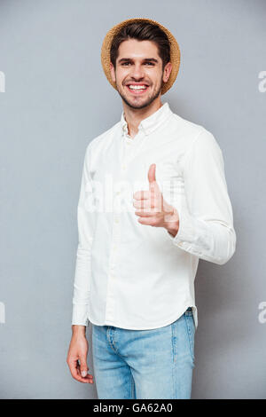 Felice Giovane Uomo in camicia bianca e hat mostra Thumbs up su sfondo grigio Foto Stock