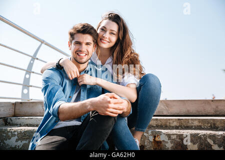 Ritratto di un sorridente coppia giovane seduto e abbracciando in riva al mare Foto Stock