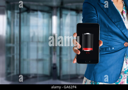 Business donna, mano tenendo un toccare telefono con la batteria scarica su uno schermo, dietro l'ingresso di un moderno edificio per uffici Foto Stock