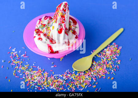 Frozen yogurt con amarena topping e rainbow spruzza su sfondo blu con giallo cucchiaio e rainbow spruzza Foto Stock