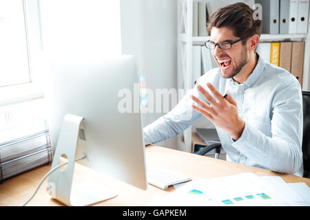 Angry mad giovane imprenditore lavora con computer e gridando in ufficio Foto Stock