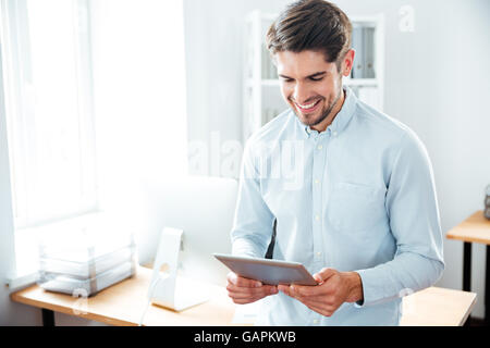 Sorridente giovane uomo in piedi e utilizzo di tablet al lavoro Foto Stock