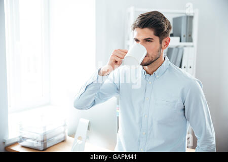 Bel giovane uomo in piedi e di bere il caffè in ufficio Foto Stock