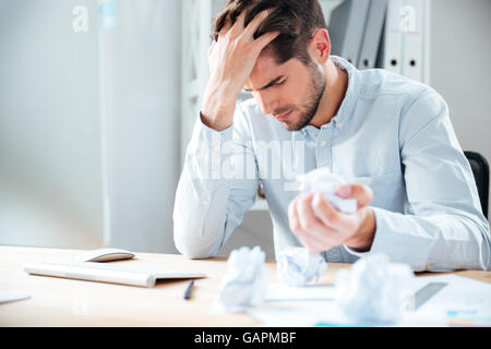 Esaurito ha sottolineato giovane uomo seduto e piegature di carta a workplace Foto Stock