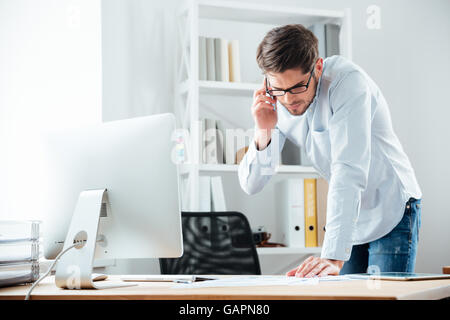 Giovani handsoome business man in piedi alla scrivania che lavora su documenti con mobile phoone in office Foto Stock