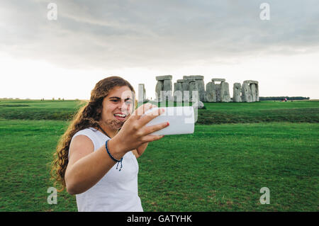 Felice giovane donna prendendo un selfie a Stonehenge un giorno nuvoloso al tramonto. Foto Stock